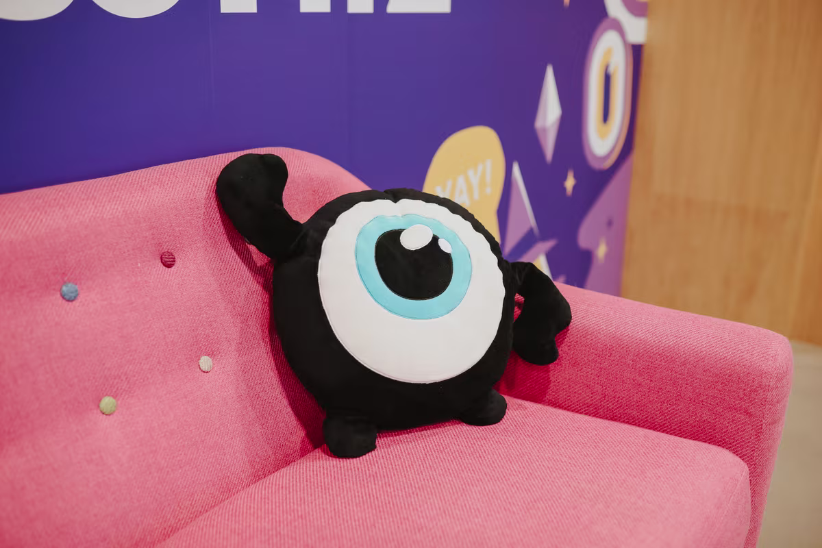 Een zwart knuffelmonster met een groot blauw oog ligt op een roze bank in een kleurrijk kantoor.