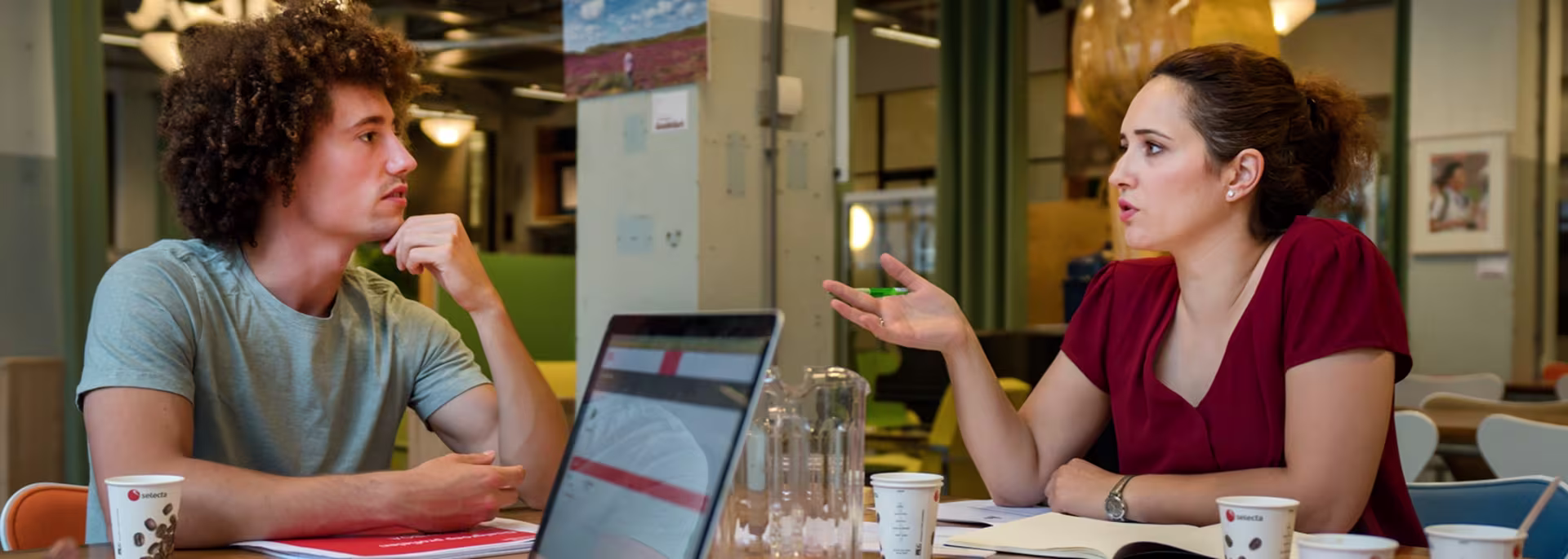 Twee vrouwen in gesprek aan tafel met laptop, koffie en water in moderne kantooromgeving.