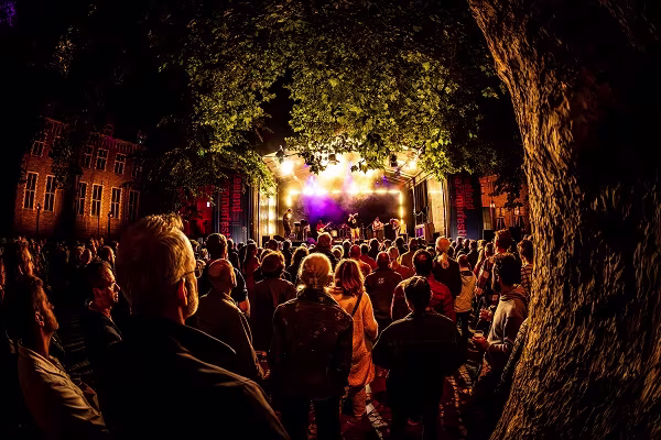 Een menigte kijkt naar een optreden op een verlicht podium tussen historische gebouwen, omringd door bomen en warme verlichting.