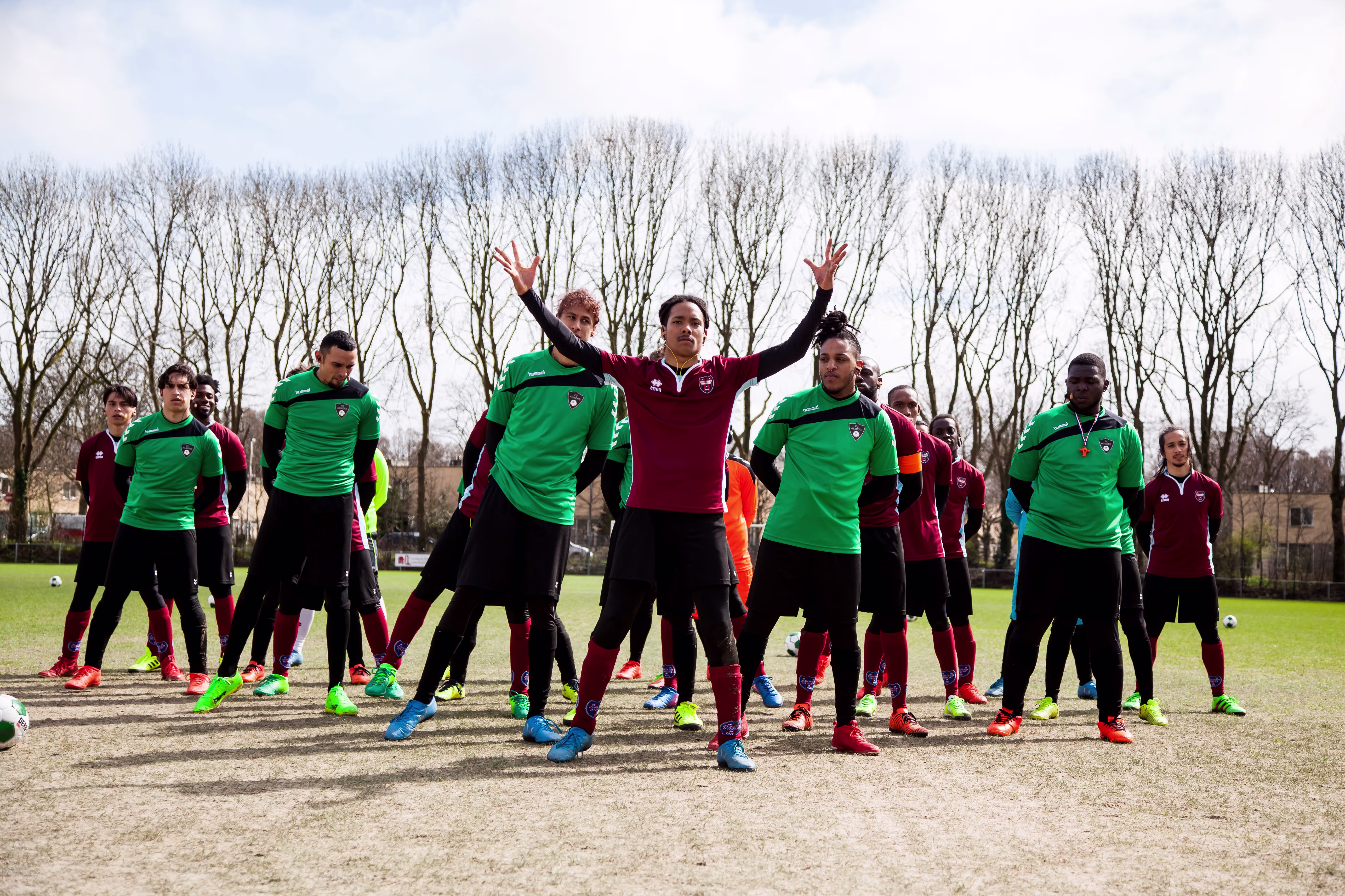 Een groep mensen in groene en rode sportkleding doet samen warming-up oefeningen op een grasveld met bomen op de achtergrond.