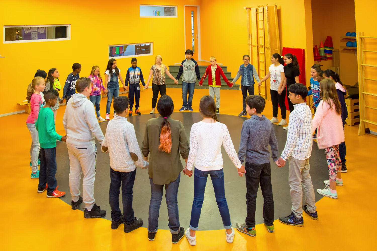Kinderen staan in een cirkel hand in hand in een klaslokaal met felgele muren tijdens een groepsactiviteit.