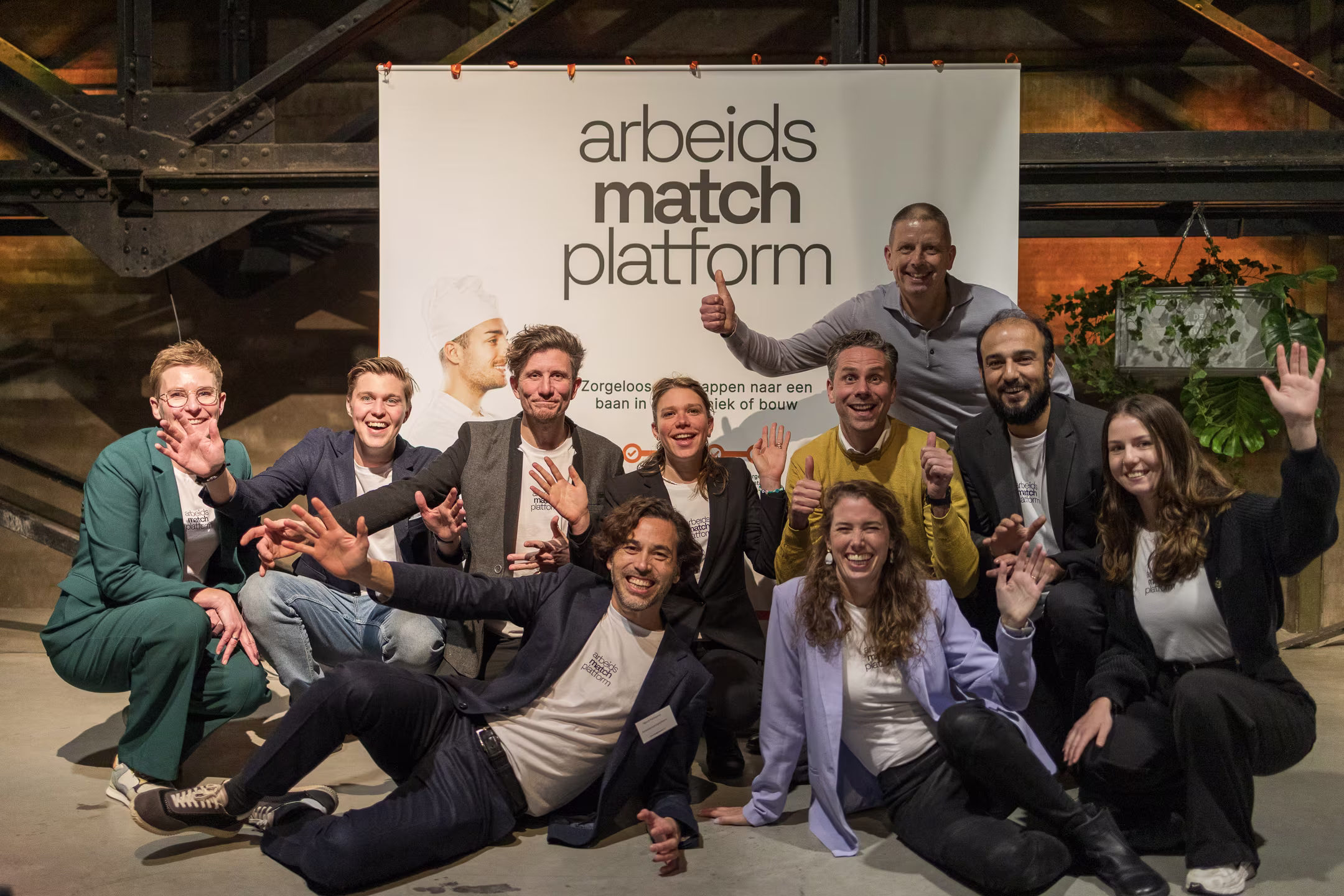 Een groep van ongeveer 10 mensen poseert vrolijk wuivend voor een banner van Arbeidsmatchplatform in een industriële ruimte.