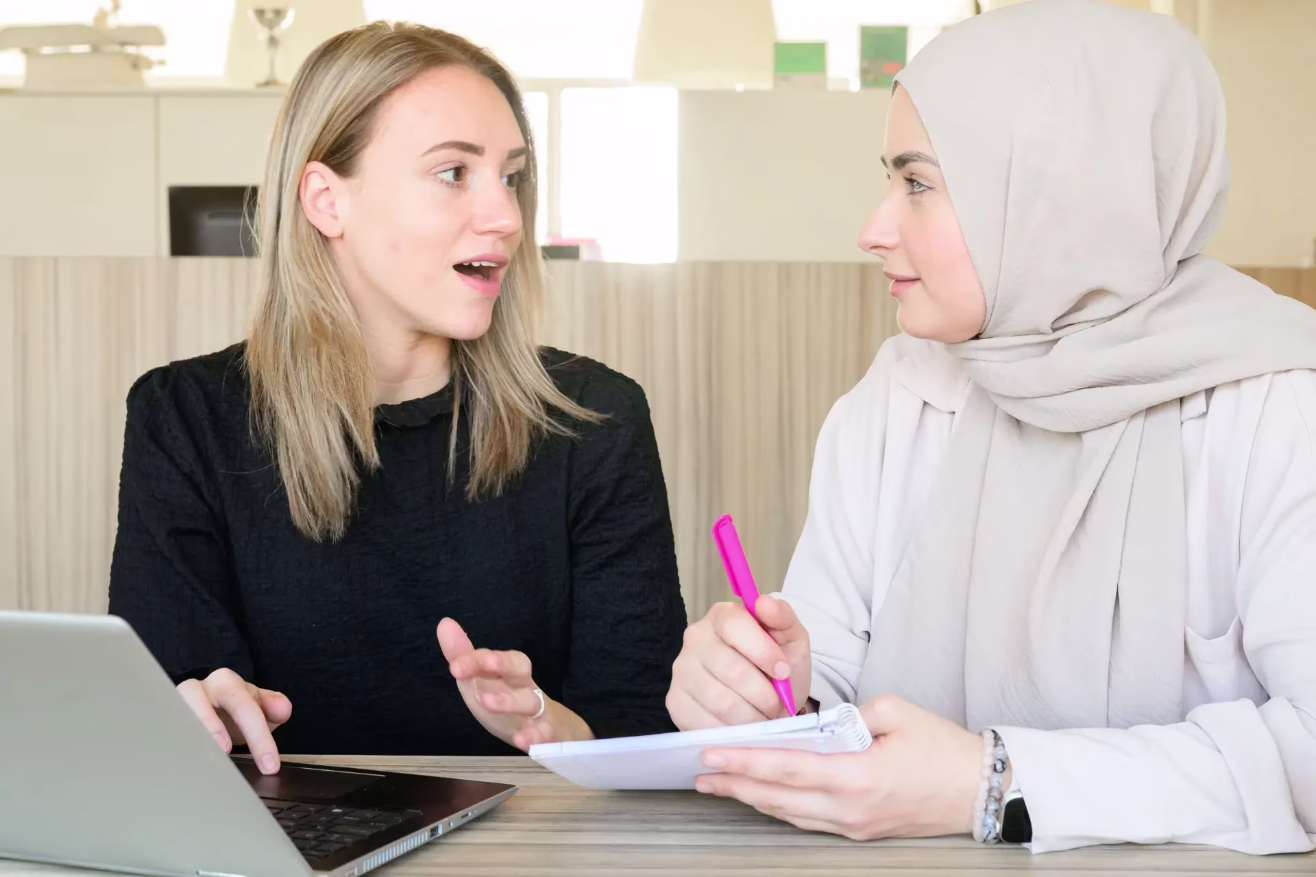 Twee vrouwelijke collega's zitten aan een bureau en voeren een gesprek tijdens het werk met laptop en notitieblok.