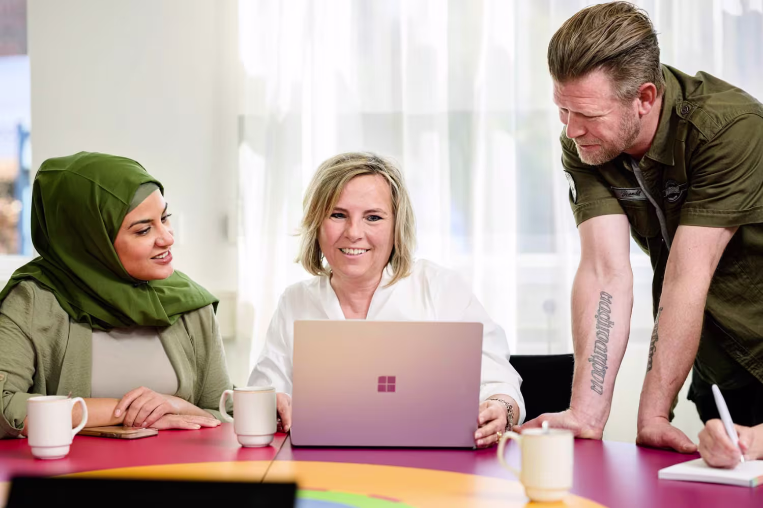 Drie collega's werken samen rond een laptop op een kleurrijke tafel in een modern kantoor.