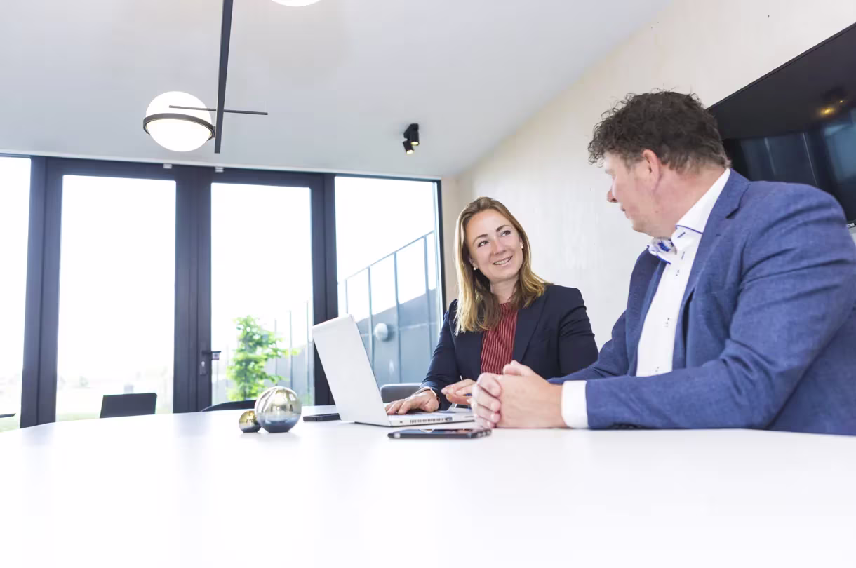 Twee professionals in pak bespreken werk achter laptop in modern kantoor met grote ramen.