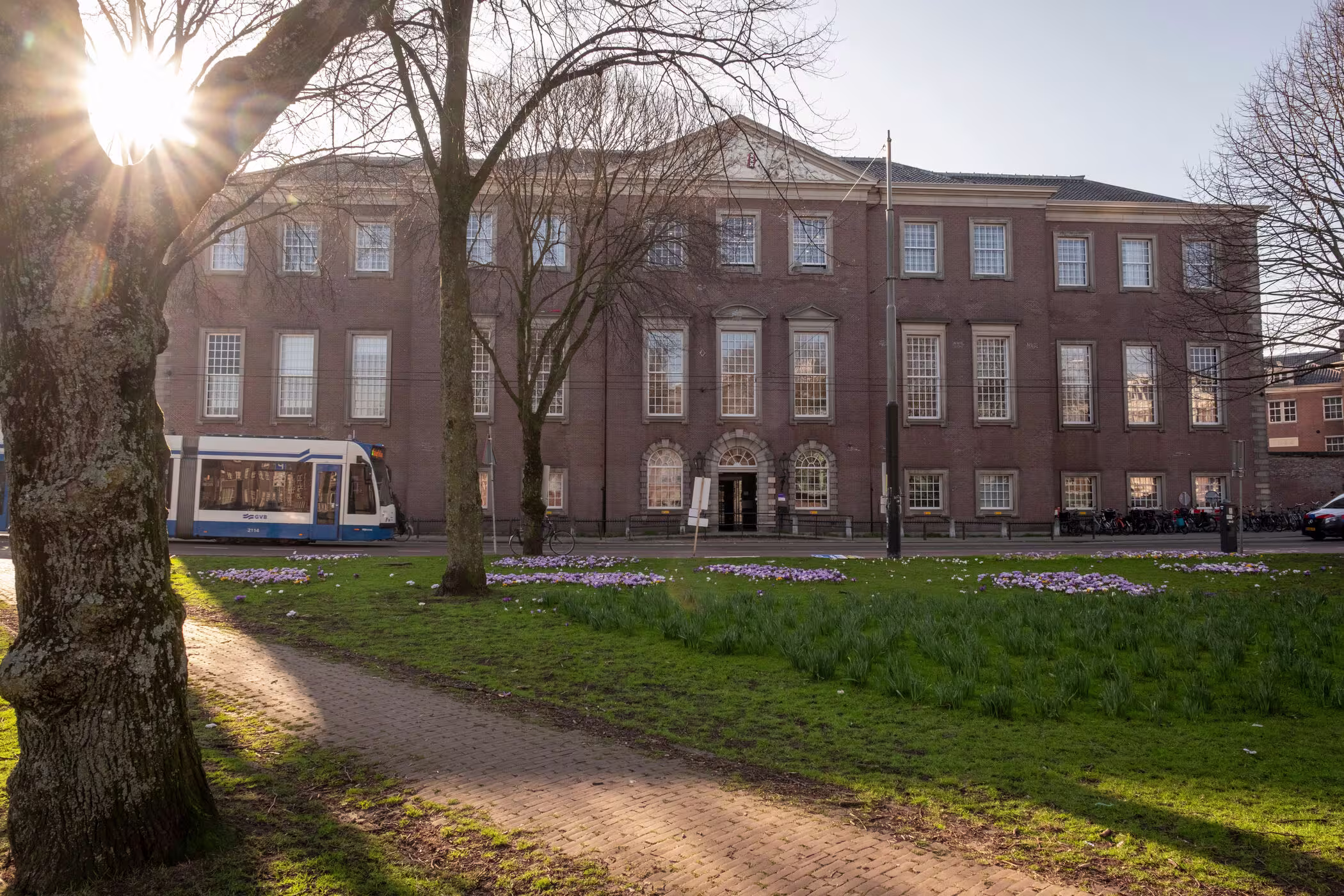 Historisch bakstenen gebouw met groene tuin ervoor, blauwe tram en zonlicht door de bomen.