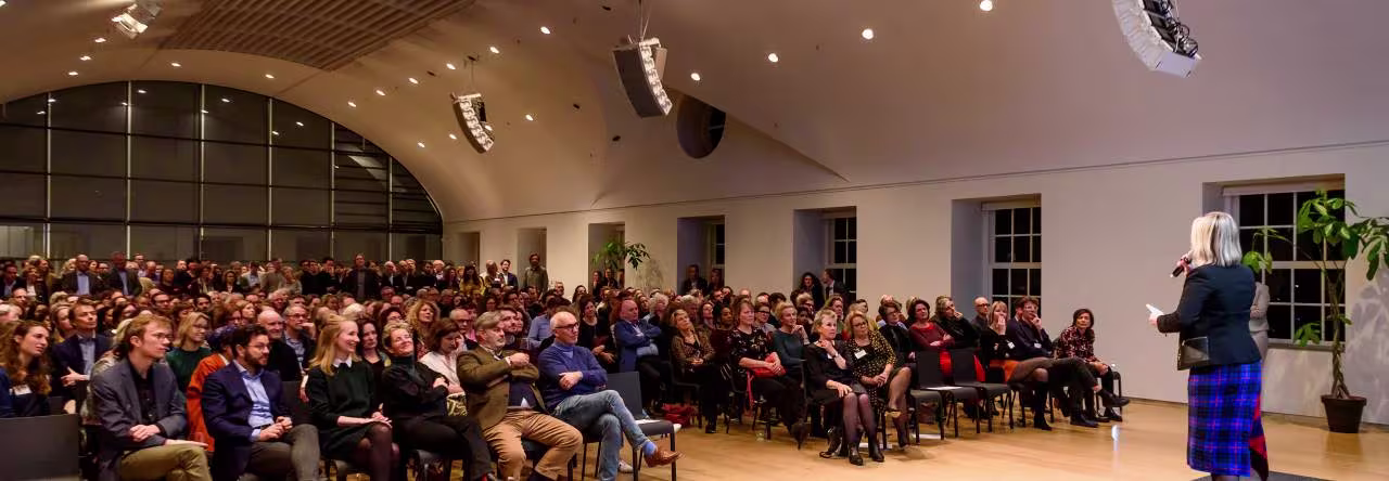 Een spreker presenteert voor een grote zaal vol publiek in een modern auditorium met grote ramen.