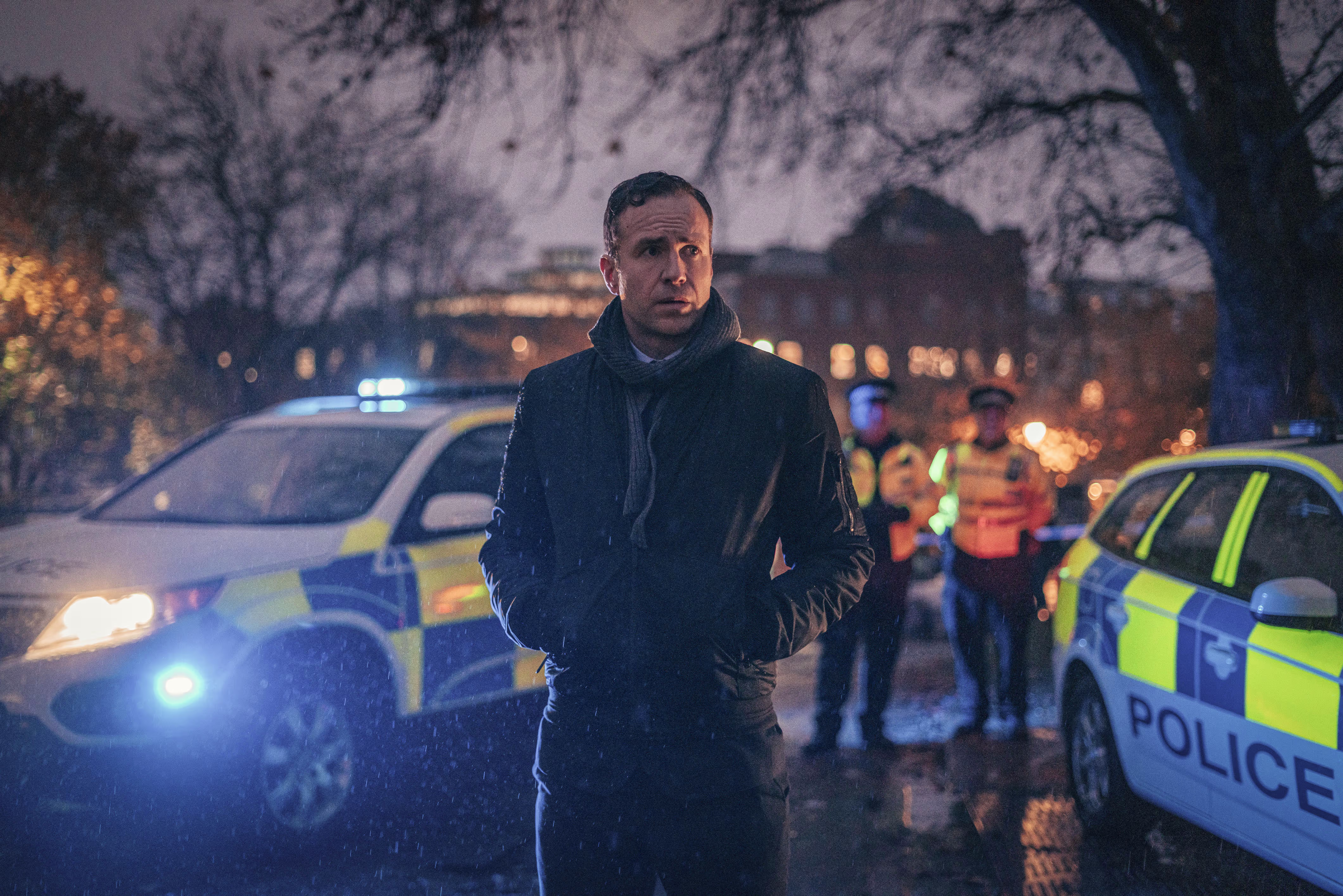 Een man in donkere jas staat bij politieauto's met blauwe zwaailichten tijdens een avondscène voor een tv-productie.