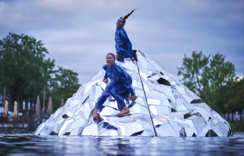 Twee performers in blauwe pakken klimmen op een witte piramide-installatie in het water tijdens een kunstvoorstelling.