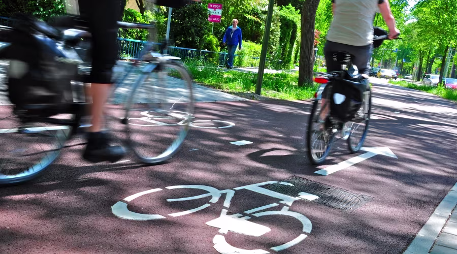 Fietsers rijden over een fietspad met fietssymbolen op de weg, omgeven door groen en bomen in een stedelijke omgeving.