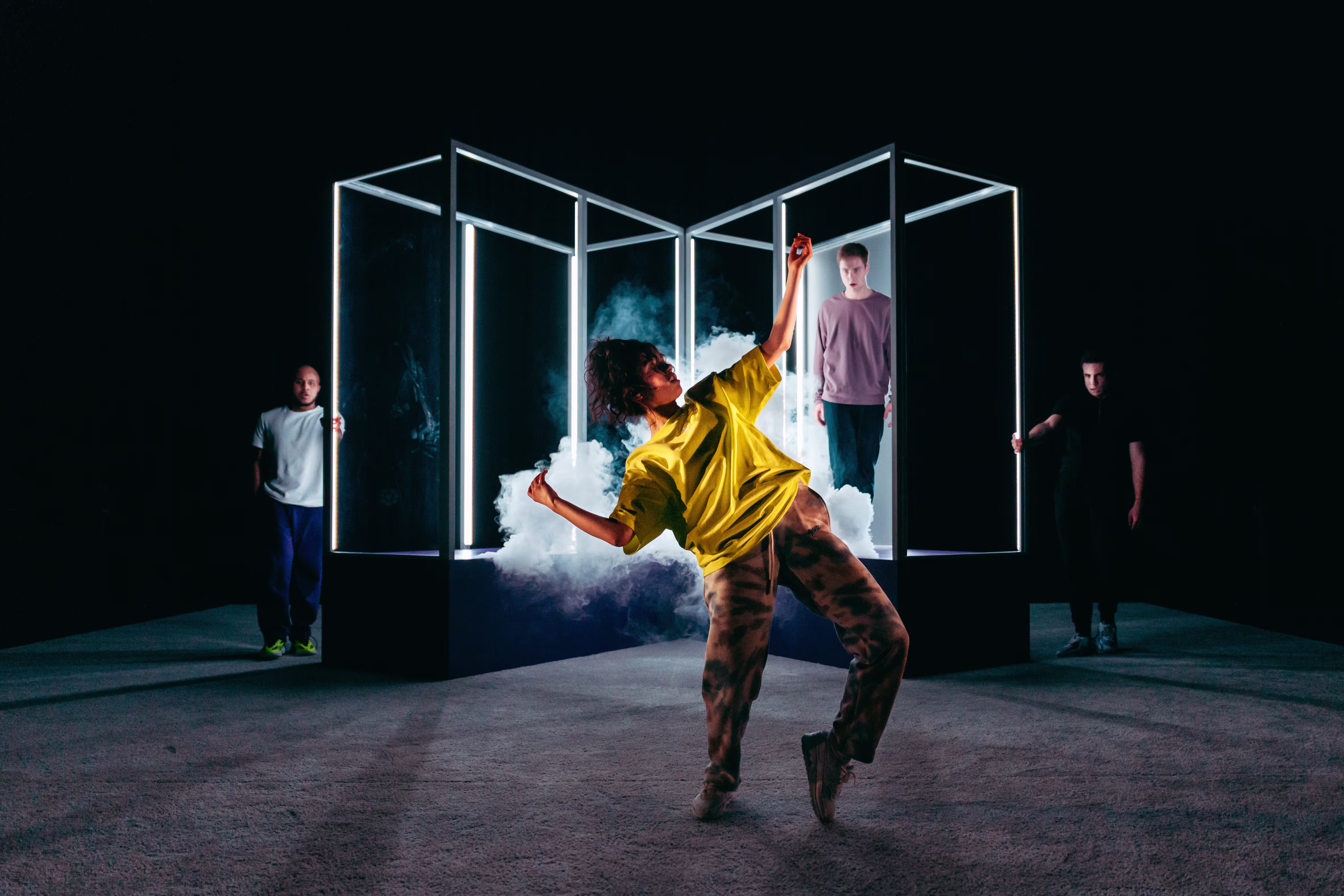 Een danser in geel shirt beweegt dynamisch voor verlichte glazen kubussen, terwijl andere performers op de achtergrond staan.