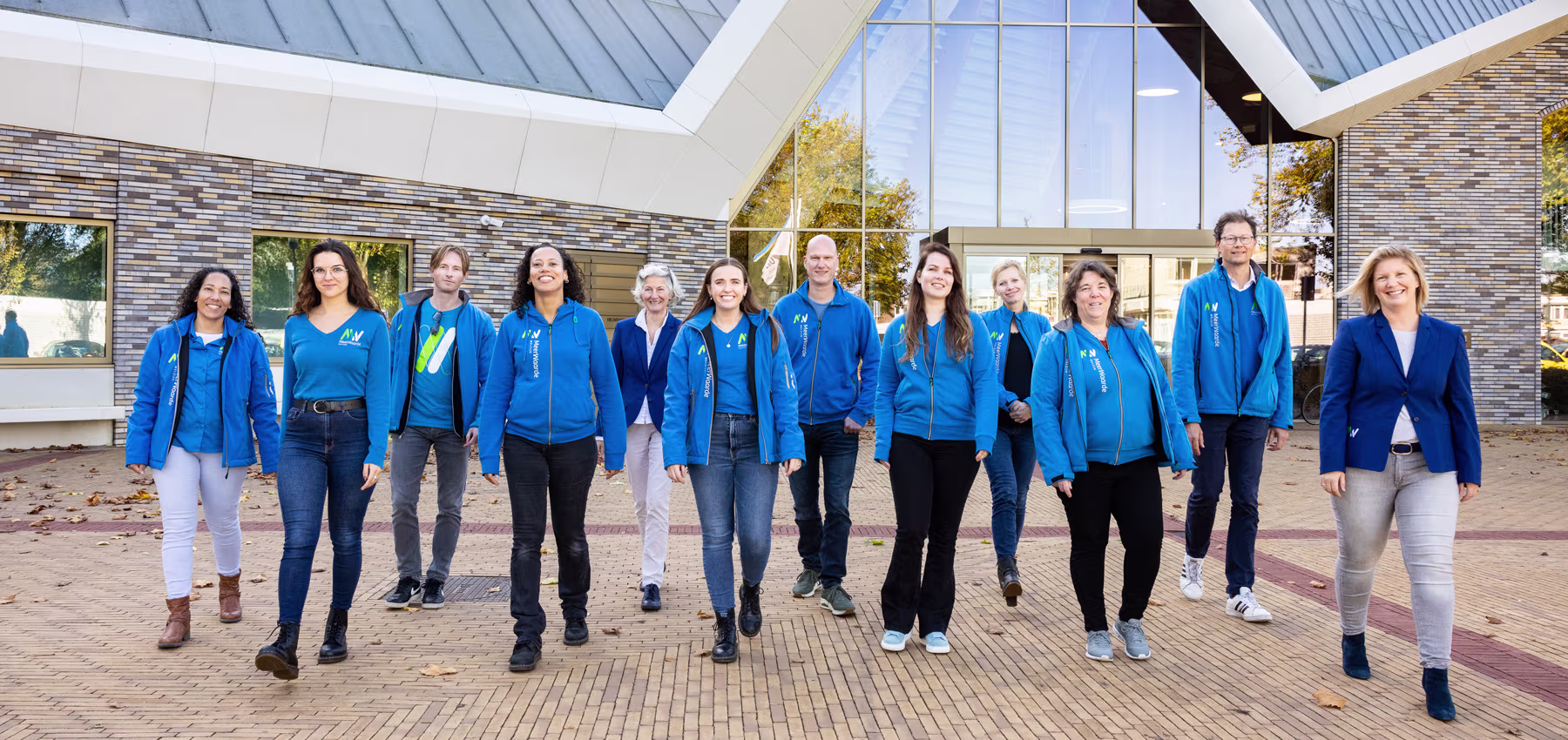 Een groep van elf medewerkers in blauwe jassen staat voor een modern gebouw met bakstenen gevel en grote ramen.
