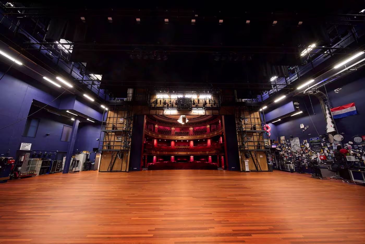 Leeg podium in theater met houten vloer, blauwe zijwanden en rood verlichte zaalruimte met balkons op de achtergrond.