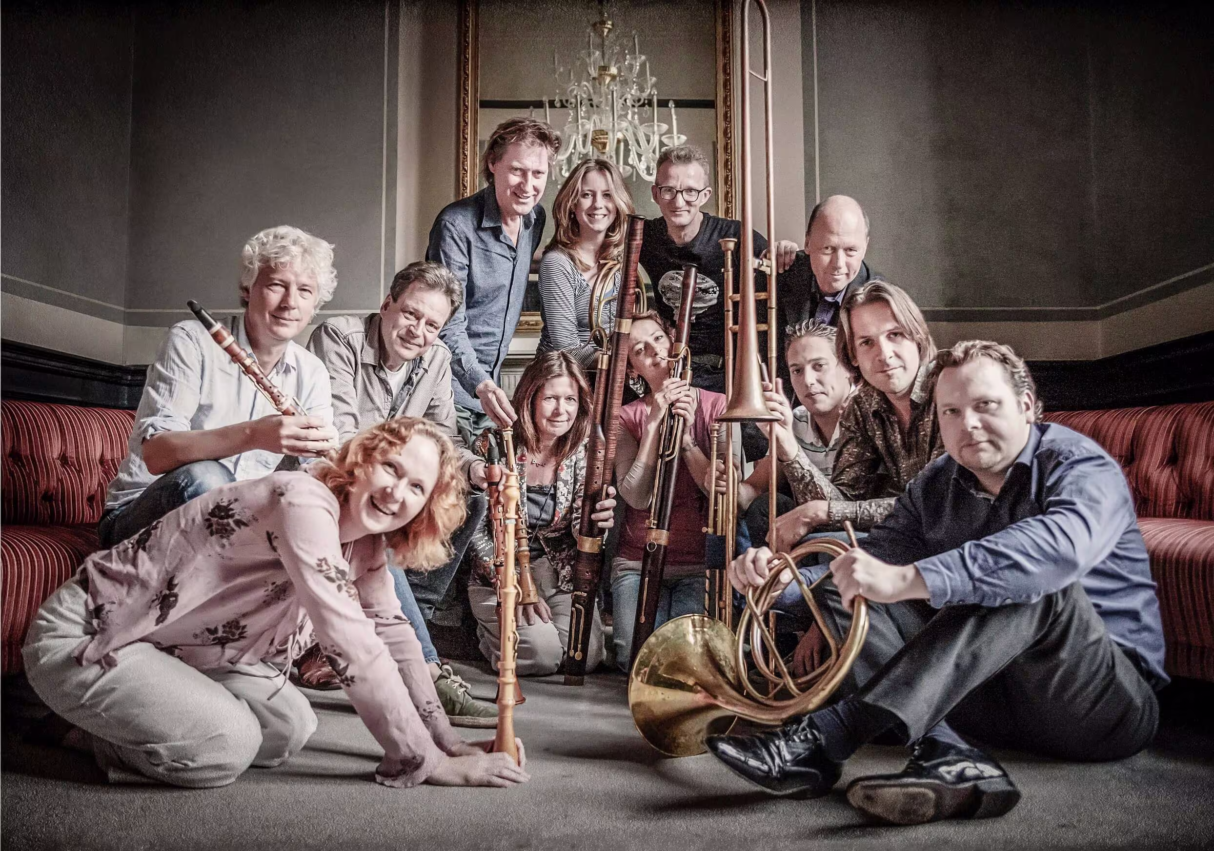 Groepsportret van het Nederlands Blazers Ensemble met blaasinstrumenten in een elegant interieur met kroonluchter.