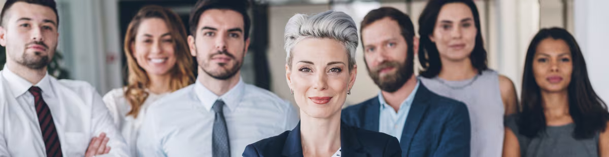 Een blonde vrouw in pak staat vooraan met zes collega's op de achtergrond in een moderne kantooromgeving.
