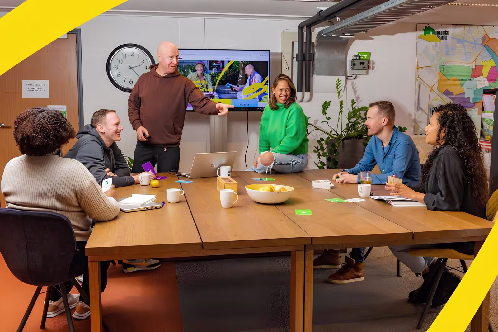 Een groep collega's in gesprek rond een houten vergadertafel in een modern kantoor met een scherm op de achtergrond.