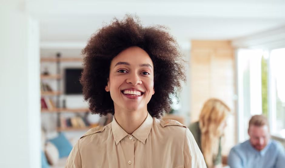 Glimlachende vrouw met krullend haar in beige blouse op een kantoor, met collega's op de achtergrond.