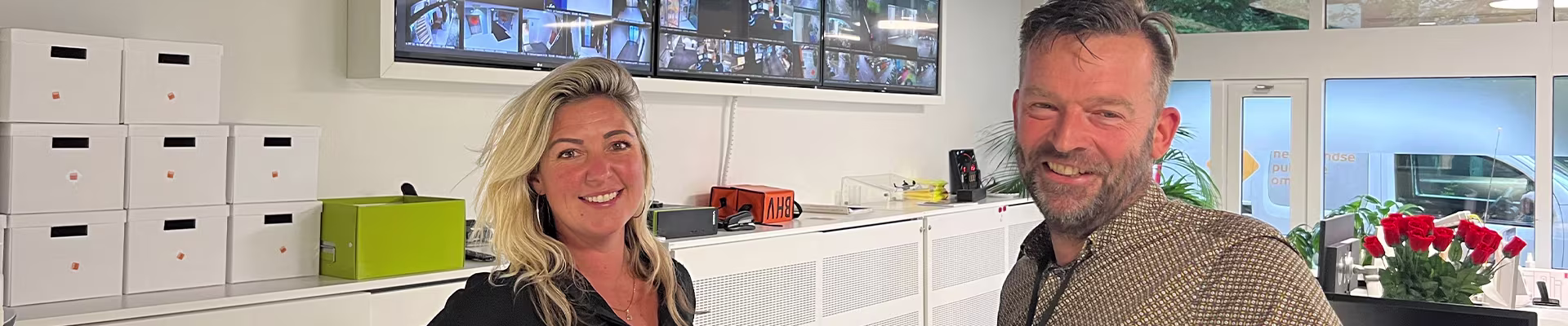Twee collega's glimlachen in een moderne kantooromgeving met monitoren en opbergboxen op de achtergrond.