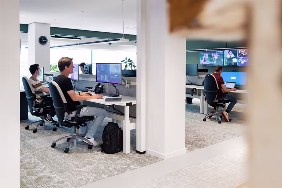 Moderne open kantoorruimte met medewerkers aan bureaus met computers en monitors, gescheiden door glazen wanden.