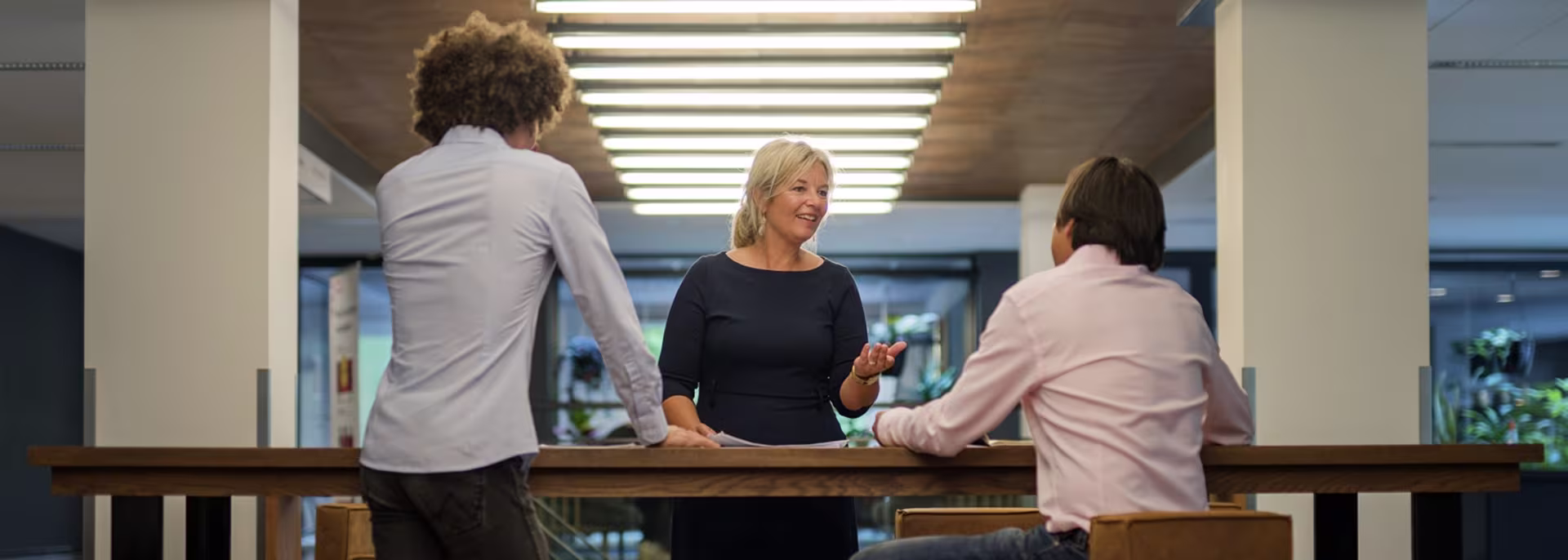 Drie collega's staan rond een houten tafel in gesprek in een modern kantoor met veel licht.