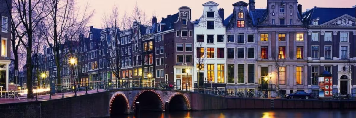 Amsterdamse grachtenpanden met verlichte ramen langs een kanaal met historische brug tijdens de avondschemering.