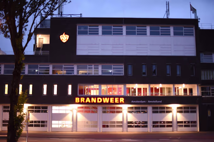 Een modern brandweergebouw van de Veiligheidsregio Amsterdam-Amstelland in de avond met verlichte ramen en garagedeuren.