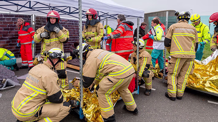 Brandweerlieden en hulpverleners in reflecterende kleding behandelen gewonden met reddingsdekens tijdens een oefening.