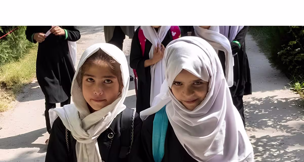 Twee jonge meisjes met witte hoofddoeken en schooluniformen lopen samen op straat, anderen op de achtergrond.