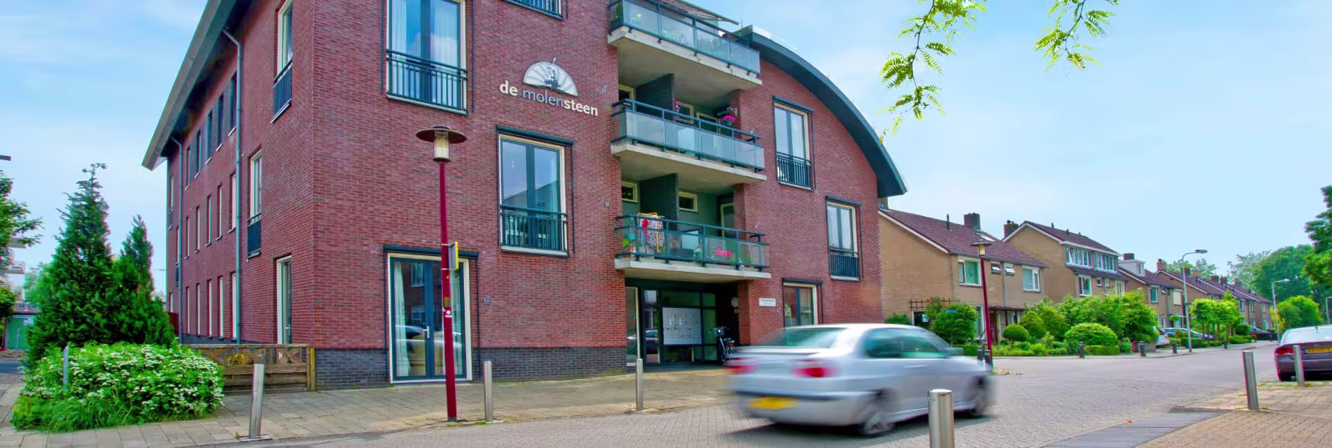 Een modern bakstenen appartementengebouw van Woondiensten Aarwoude met balkons en een passerende auto op straat.