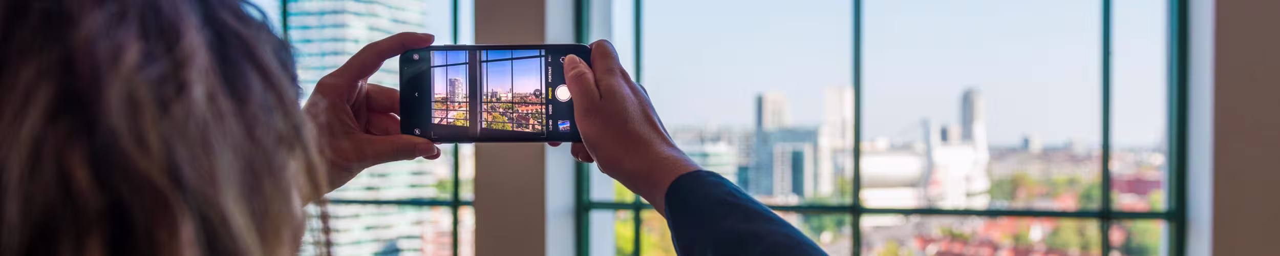 Een persoon fotografeert de skyline van Eindhoven met een smartphone vanuit een kantoorgebouw met grote ramen.