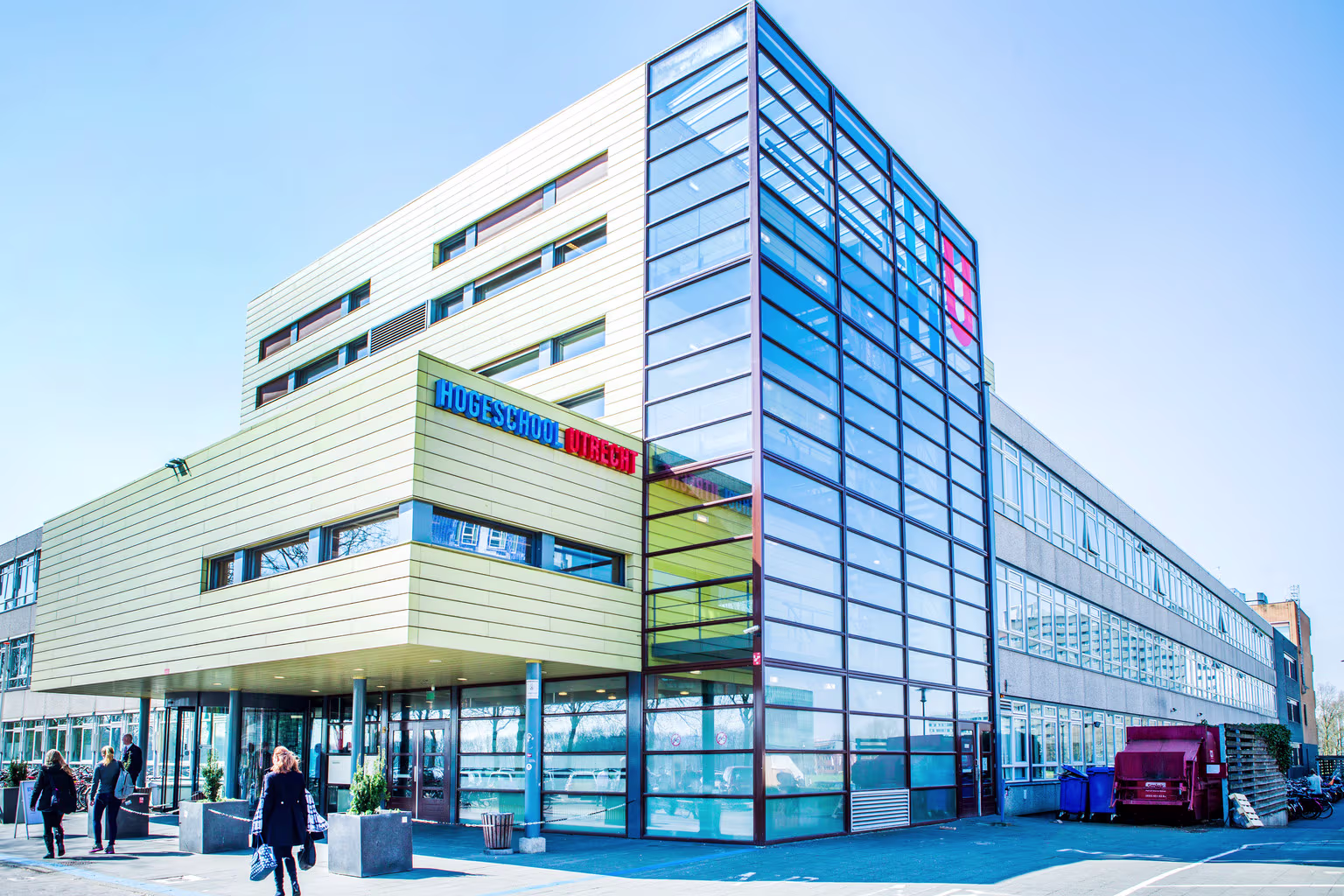 Modern glazen gebouw van Hogeschool Utrecht met studenten en bezoekers op het plein ervoor onder een blauwe hemel.