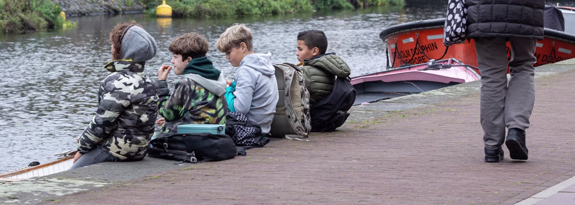 Een groep kinderen zit samen aan de waterkant bij boten, terwijl een volwassene in waders dichtbij staat.