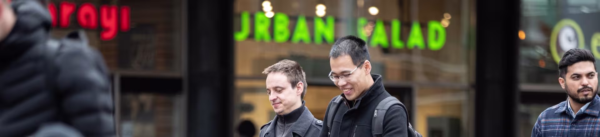 Drie mannen lopen lachend door een winkelstraat met winkels op de achtergrond, een informele werksfeer.