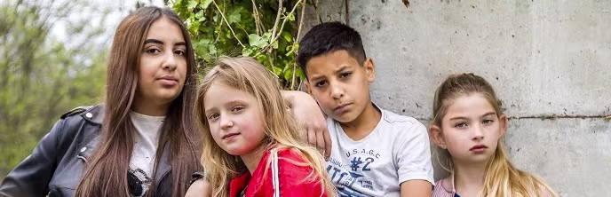 Vier kinderen van verschillende leeftijden zitten samen tegen een betonnen muur, ze kijken vriendelijk in de camera.