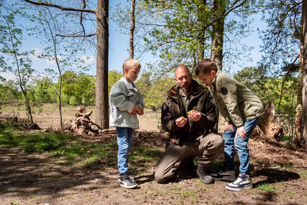 Een medewerker van GNR toont twee kinderen iets in de natuur tussen de bomen op een zonnige dag.
