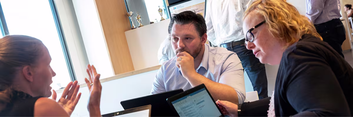 Drie collega's in gesprek rond een laptop op kantoor, waarbij één persoon iets uitlegt met handgebaren.