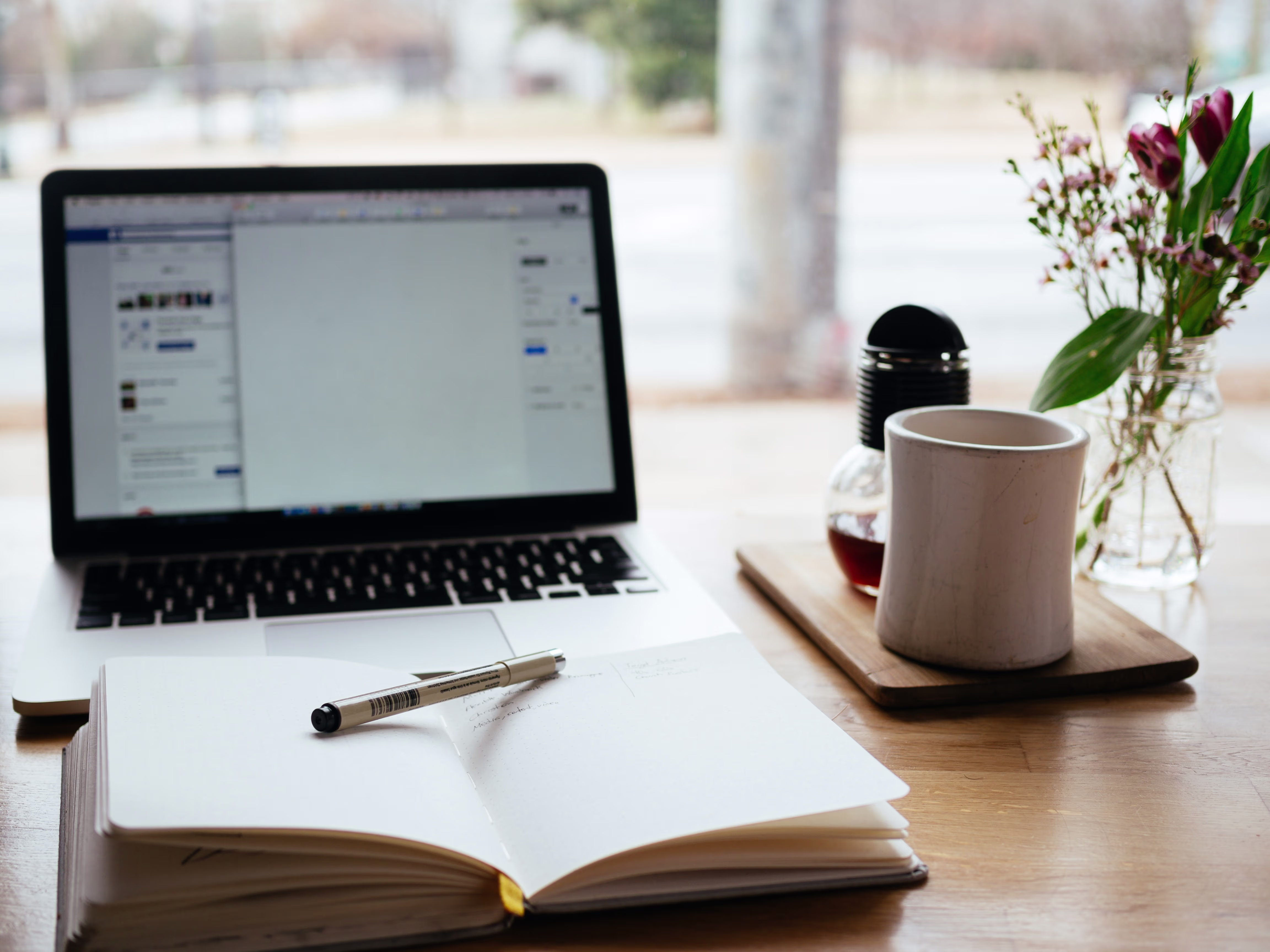Een laptop, opengeslagen notitieboek met pen, koffiemok op houten dienblad en bloemen in vaas op een houten bureau.