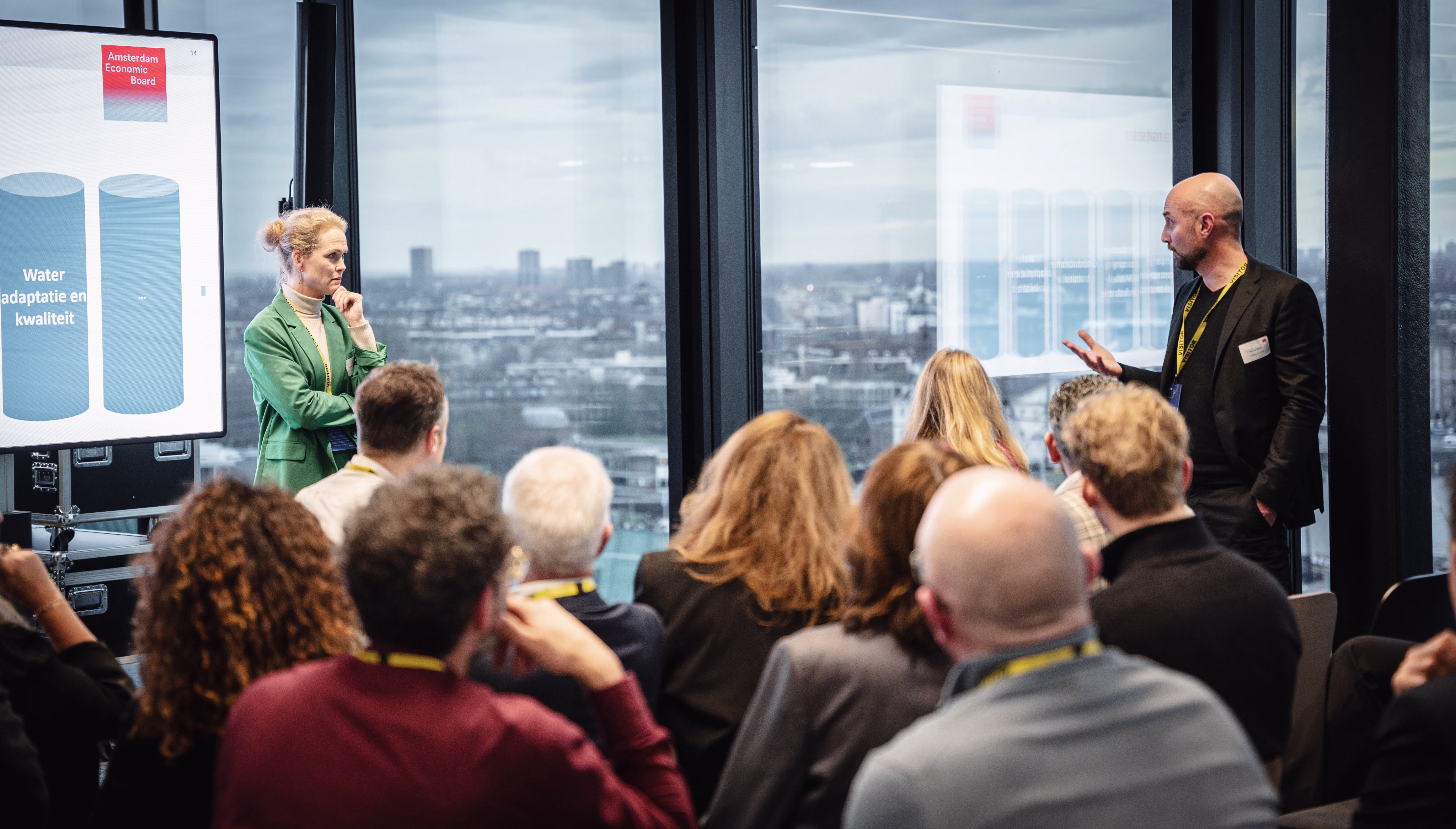 Presentatie in moderne vergaderruimte met uitzicht over de stad, spreker en publiek luisteren aandachtig naar de uiteenzetting.