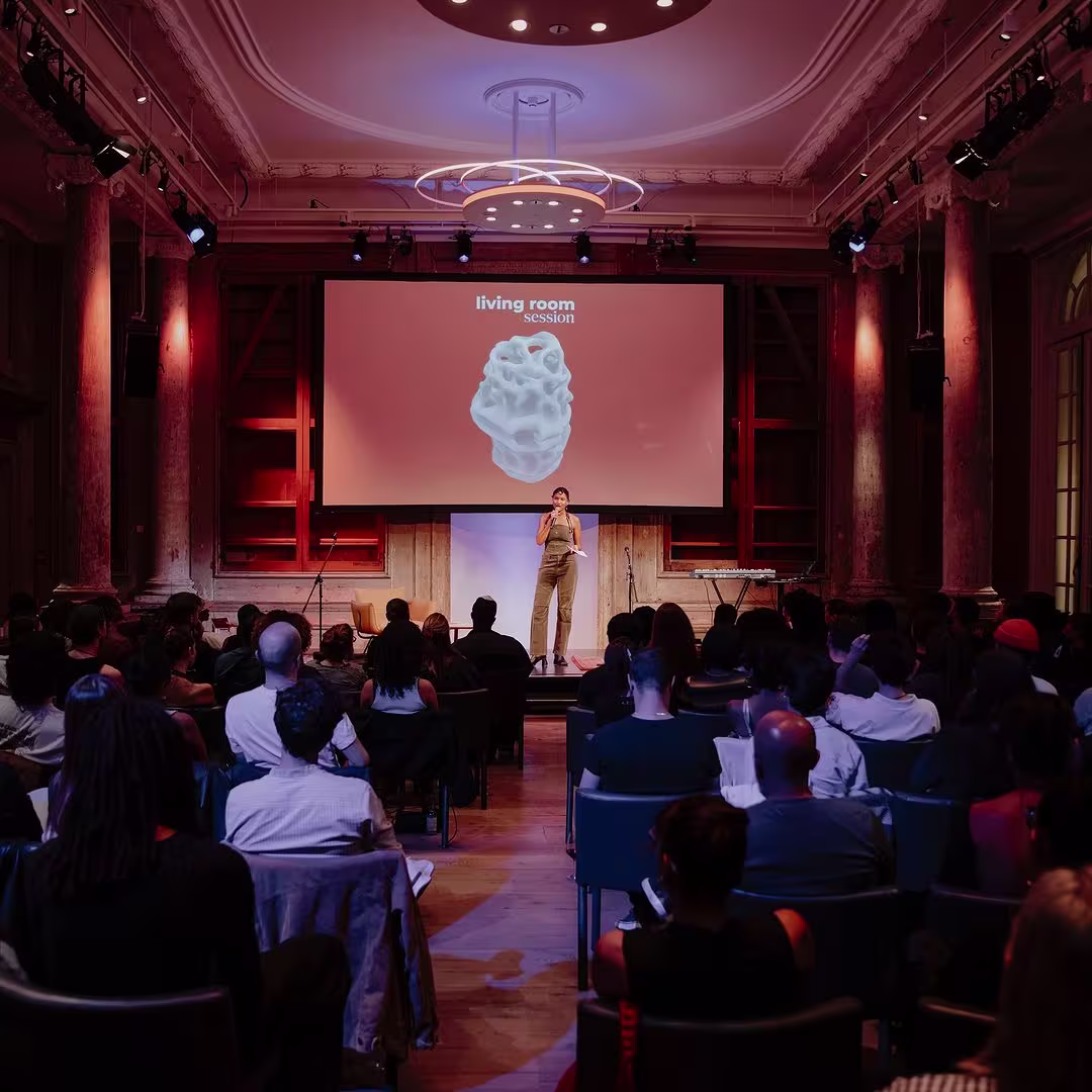Een spreker presenteert op het podium voor een publiek in een historische zaal met rode verlichting en grote projectiescherm.