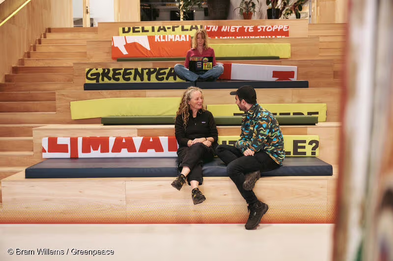Drie mensen zitten op houten trappen met Greenpeace klimaat-banners erop in een modern kantoorgebouw.