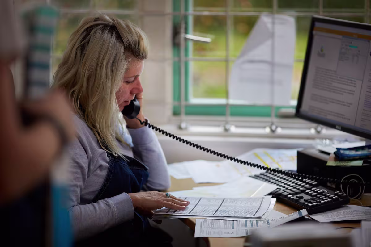 Blonde vrouw belt aan bureau met computer en papieren, kantooromgeving met raam op achtergrond.