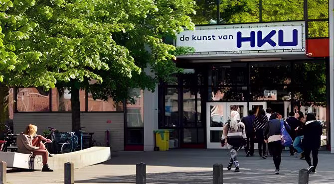 Studenten lopen voor het HKU-gebouw met groene bomen en een persoon zittend op een bankje in de voorgrond.