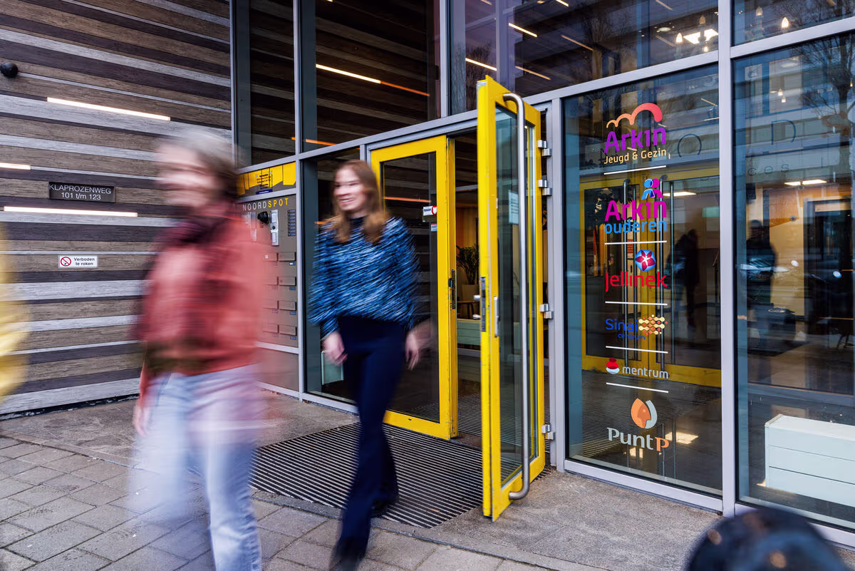 Mensen lopen door de gele toegangsdeur van een modern glazen gebouw met het Arkin logo op de gevel.