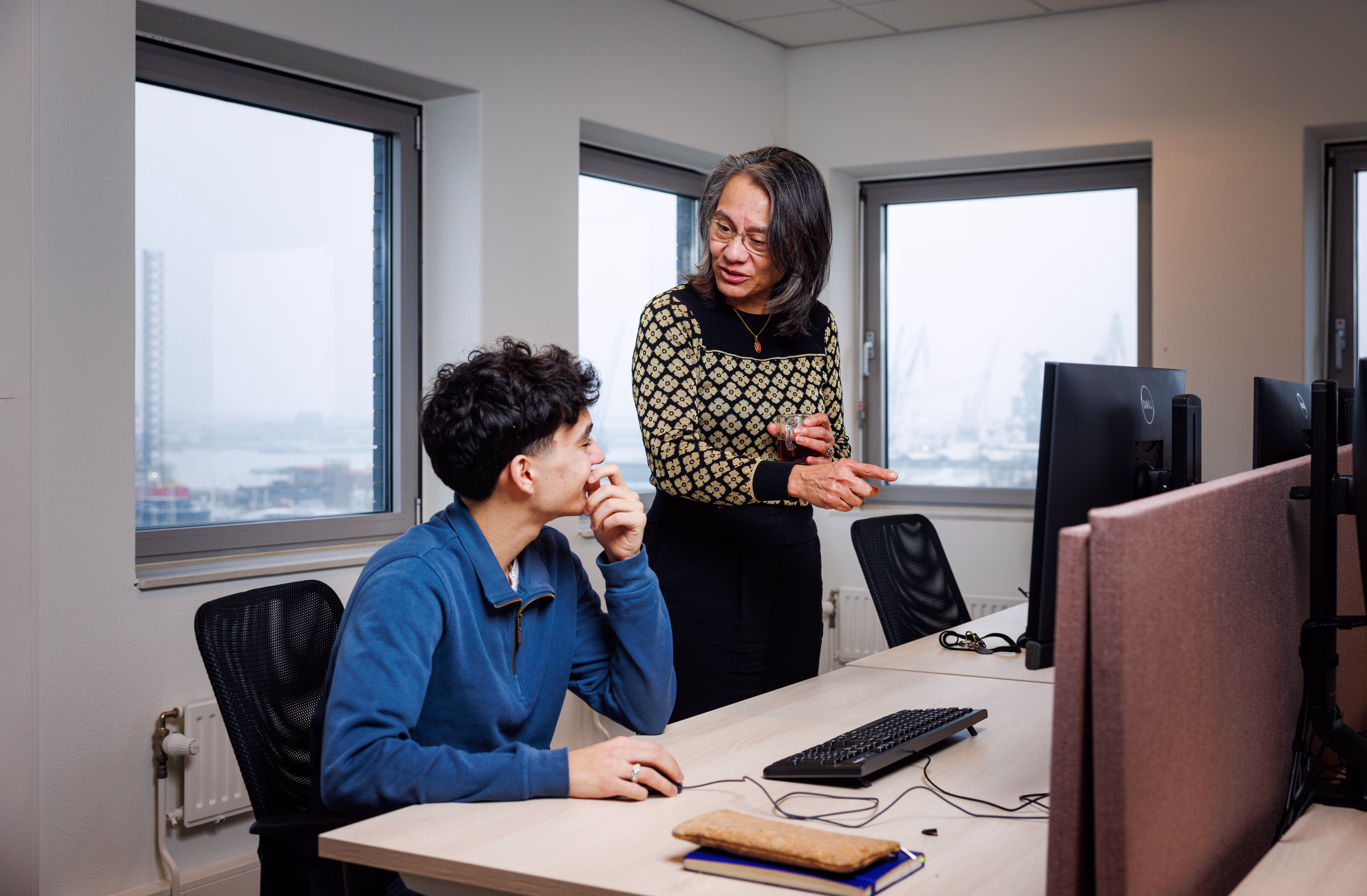 Twee collega's werken samen aan een computer in een modern kantoor met grote ramen en uitzicht op de stad.