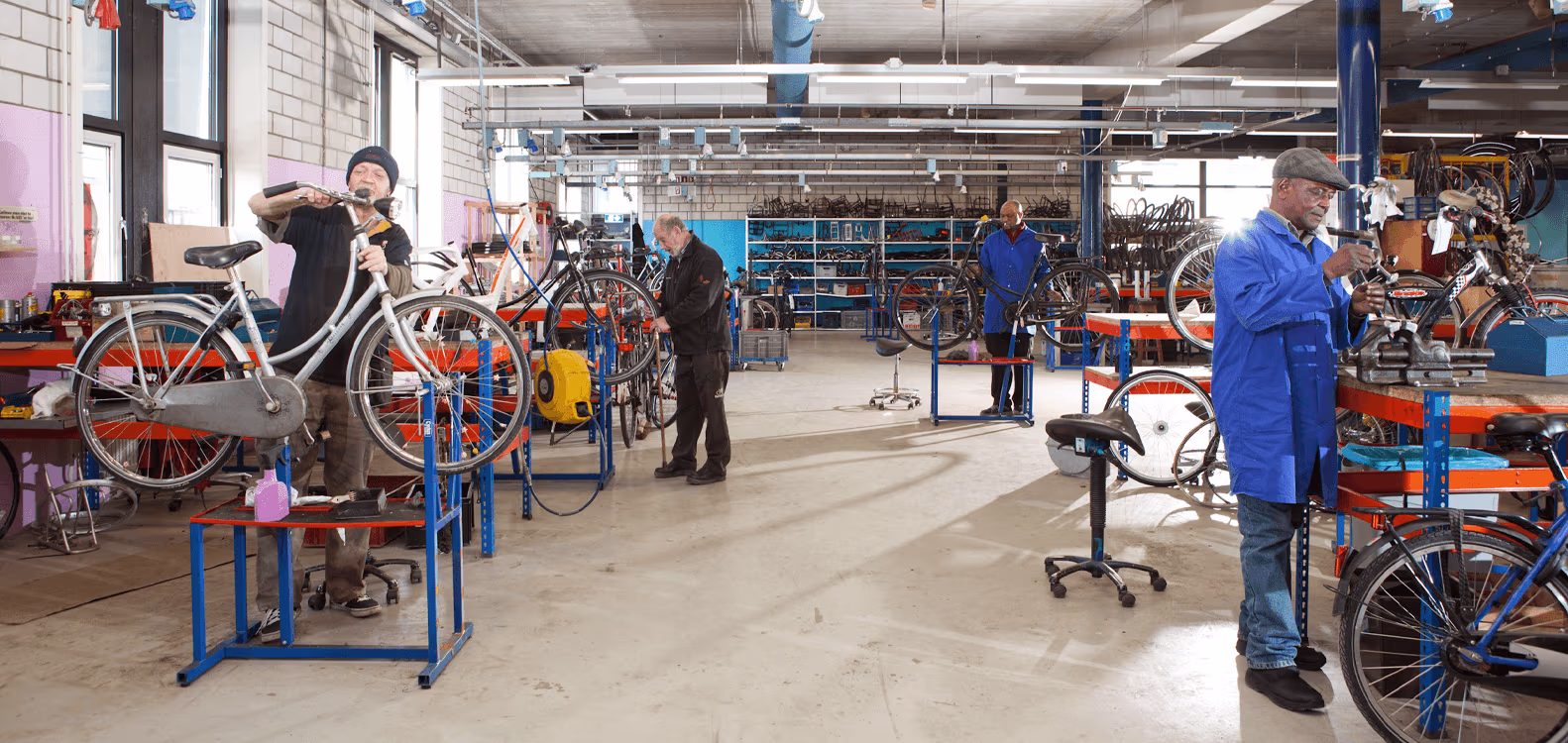 Medewerkers in blauwe werkkleding repareren fietsen in een grote werkplaats met veel fietsen op werkbanken.