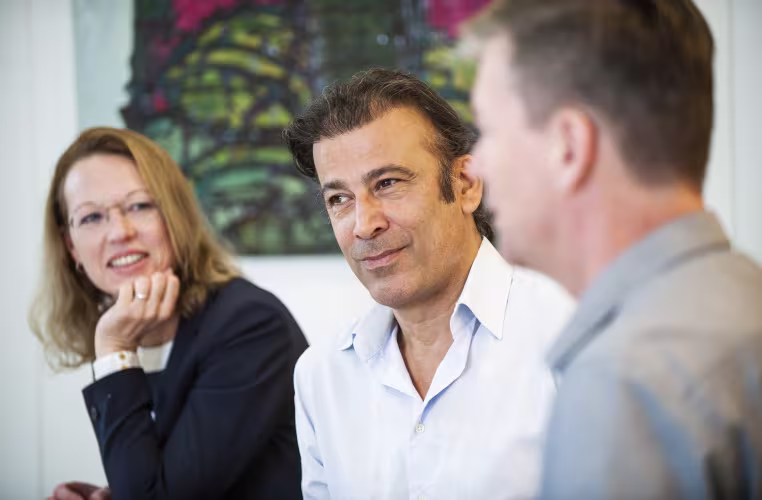 Drie collega's in gesprek op kantoor, een vrouw en twee mannen glimlachen tijdens een informele werkoverleg.