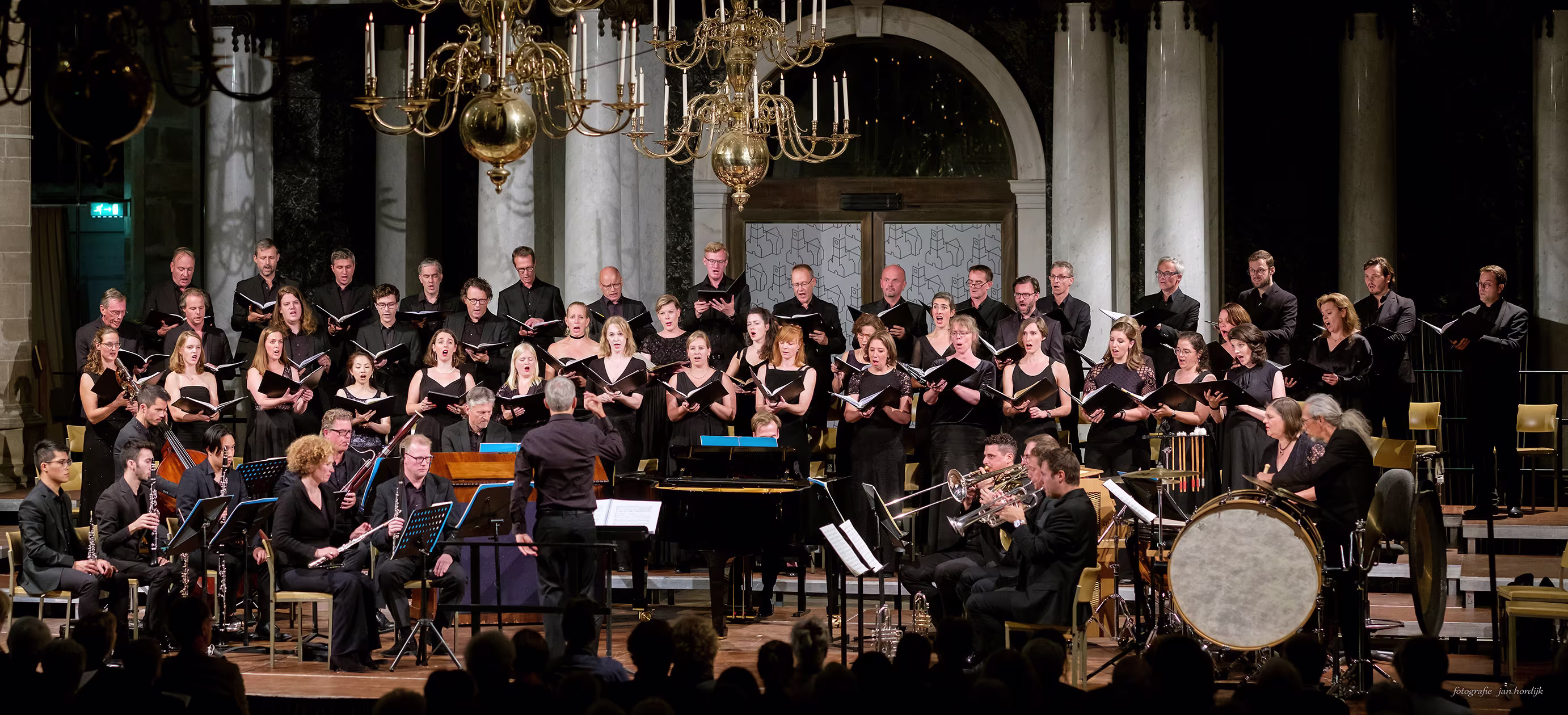 Een koor in zwarte kleding treedt op in een kerk met orkestbegeleiding en een dirigent voor het publiek.