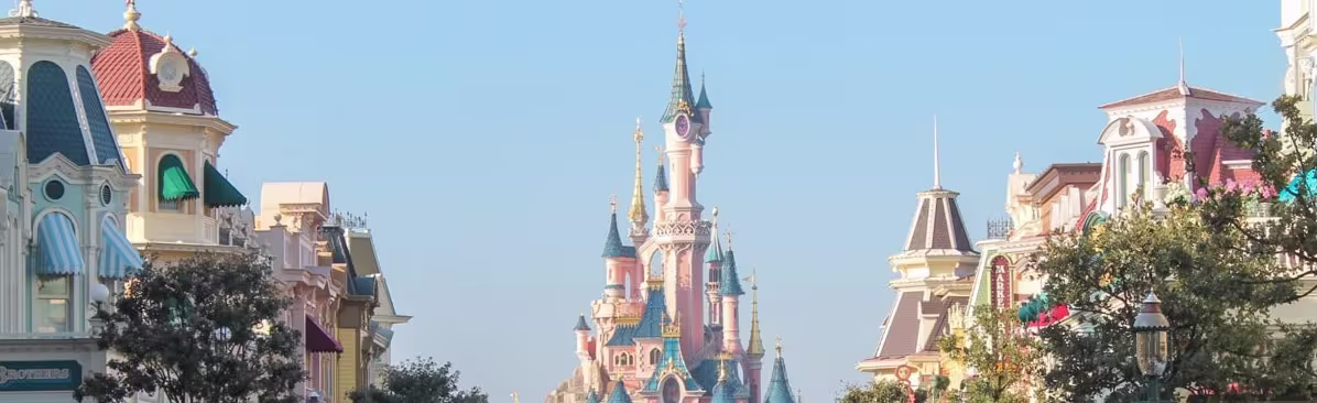 Kleurrijk Disney-kasteel met roze en blauwe torens, omringd door sprookjesachtige gebouwen en groen in een themapark.