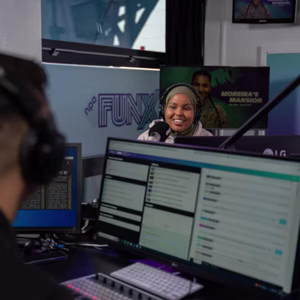 Een vrouw met hoofddoek en koptelefoon presenteert in een radiostudio van FunX, omringd door computerschermen.