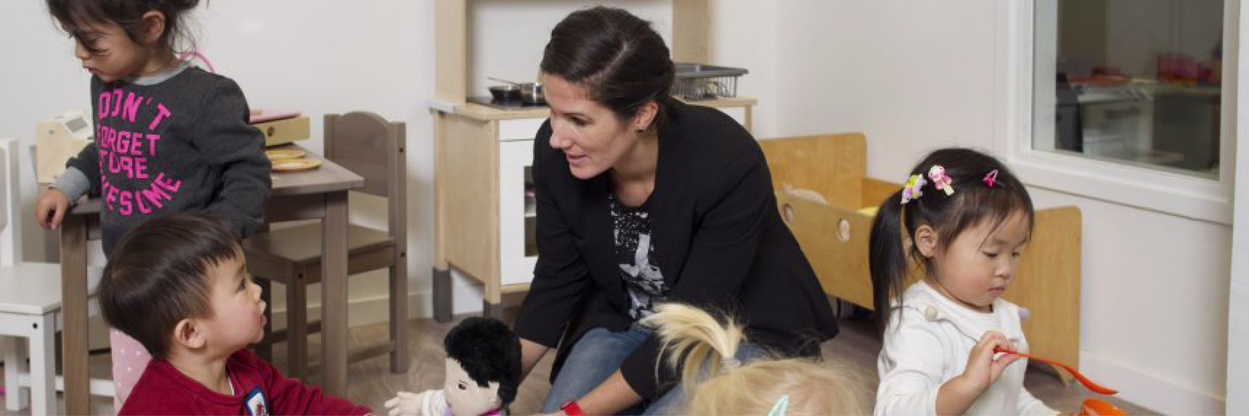 Een leidster zit op de grond en speelt met kinderen in een kinderdagverblijf met houten stoeltjes en tafels op de achtergrond.